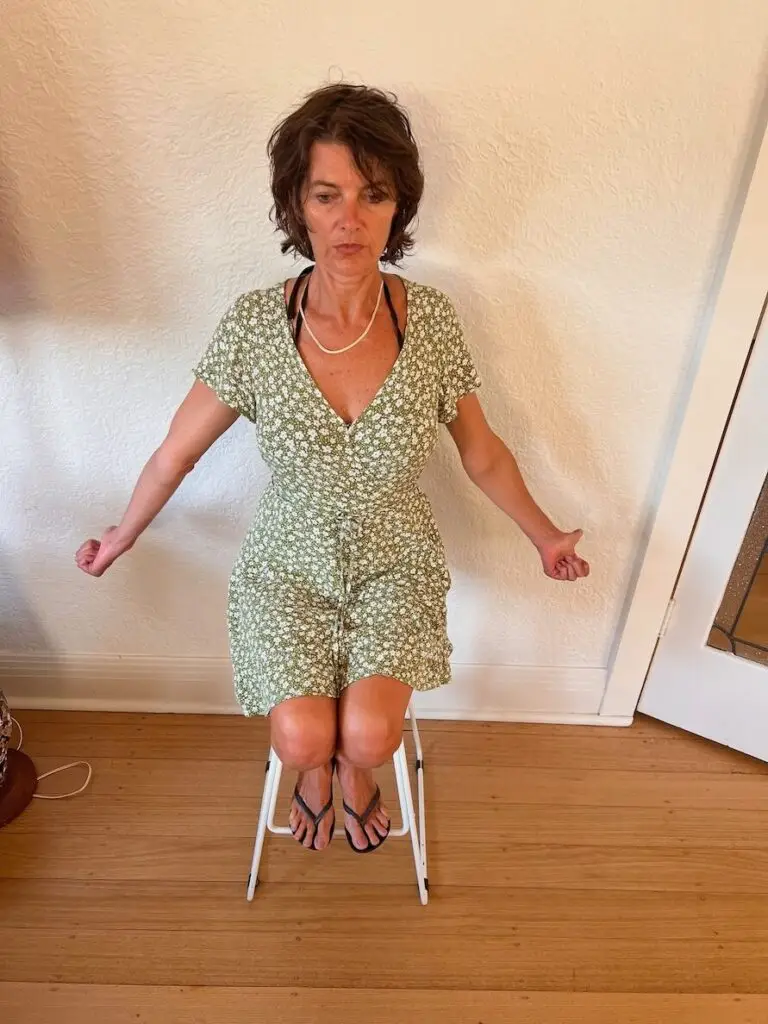 woman viewed front on sitting on a stool adopting Bruggers relief position whilst externally rotating her arms and extending her shoulders